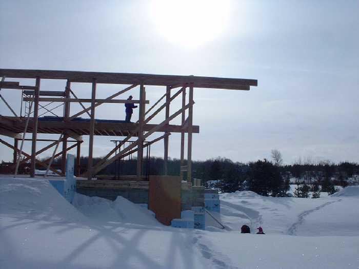 Looking south through the post and beam construction. Leigh surveys the second floor while Ian and Claire play in the snow to the left. The sun shines down through the site.